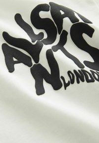 Bílá tkanina s tučnými, nepravidelnými černými nápisy "ALSA NTS LONDON." Textura vypadá hladce a mírně pružně.