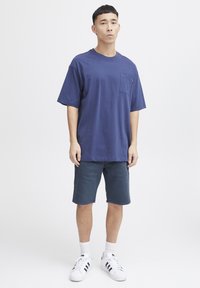 Tampak navy modra bombažna majica s žepom na prsih. Oblečena z temnimi kratkimi hlačami in belimi športnimi copati z črnimi črtami. Enostavna ozadje.