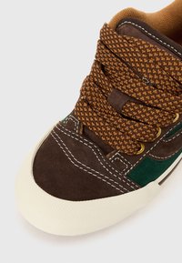 Hnědé a zelené semišové tenisky s tlustými hnědými šněrovacími tkaničkami, bílým švem a texturovaným krémově bílým gumovým špičkou.