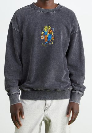 Dunkelgraues, ausgebleichtes Sweatshirt mit farbenfrohen, bestickten Sonnen- und Mondfiguren, die sich in der Mitte der Brust an den Händen halten, getragen von einer Person mit hellen Hosen.
