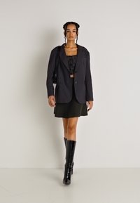 Čierny sakový blejzer cez čierny krátky top, skombinovaný so zelenou mini sukňou a čiernymi vysokými čižmami. Hladká tkanina a štruktúrovaný dizajn.