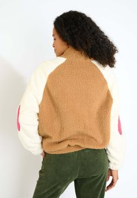 Giacca in pile marrone e crema con dettagli rosa, colletto alto e tessuto testurizzato; presenta una vestibilità rilassata e maniche di colore contrastante.