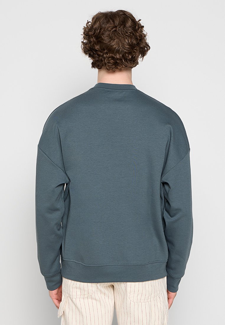 Eine person mit lockigem haar, die mit dem Rücken zur Kamera steht, trägt einen schlichten dunkelgrauen Sweater und hellcremefarbene Hose mit dünnen roten senkrechten Streifen.