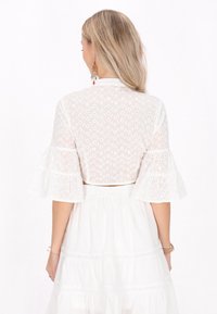 Blusa bianca corta con maniche a campana e ricami floreali. La vista posteriore mostra un tessuto leggero con un motivo aperto delicato.