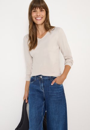 Hellbeige Langarmshirt mit V-Ausschnitt, kombiniert mit blauen weiten Jeans. Es verfügt über eine Brusttasche und einen Knopfverschluss. Sanfte Textur, lässiger Stil.