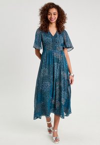 Robe imprimée florale turquoise avec des manches courtes et évasées, un décolleté en V et une taille froncée, associée à des talons fins argentés.