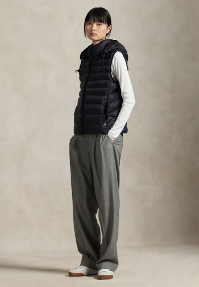 Un mannequin se tient devant un fond beige uni, portant un gilet matelassé noir par-dessus un haut blanc, un pantalon gris ample et des baskets blanches.