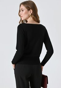 Maglione nero a maniche lunghe con scollo ampio, fondo aderente e dettagli con bottoni sulle maniche, abbinato a pantaloni neri sartoriali e una borsa rossa.