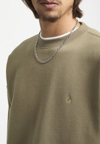 Sweatshirt vert olive avec un col côtelé, présentant un petit logo doré sur la poitrine. Associé à un collier en chaîne argentée.