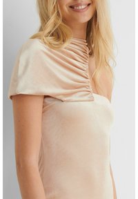 Robe en velours beige avec un design drapé sur une épaule. Le tissu a une texture douce, avec des détails fluides le long du corsage.