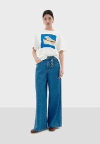 Witte t-shirt met een blauw en beige grafische print, gecombineerd met blauwe wijde spijkerbroeken. Sleutelhanger bevestigd aan de tailleband, bruine schoenen.