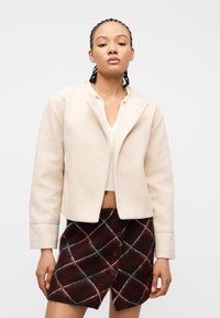 Γυναίκα που φοράει μια κρεμ κοντή ζακέτα πάνω από λευκό τοπ και μια κουμπωτή καρό φούστα σε σκούρο καφέ, στέκεται μπροστά σε απλό φόντο.