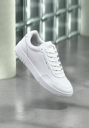 Zapatilla blanca de caña baja con cordones, logotipo de Tommy Hilfiger en la lengüeta y el lateral, flotando sobre un suelo de hormigón gris en un ambiente interior moderno.