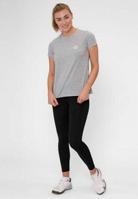Vrouw staat en glimlacht, gekleed in een lichtgrijze t-shirt, zwarte leggings en witte sportschoenen tegen een effen achtergrond.
