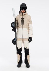Giacca da snowboard beige e nera con numerose tasche con zip, abbinata a pantaloni coordinati con grandi tasche cargo e attrezzatura da snowboard.