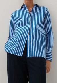 Modrá a bílá pruhovaná košile s dlouhým rukávem, která má špičatý límec a mírně uvolněný střih, v kombinaci s tmavými kalhotami.