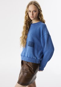 Blauwe sweatshirt met ronde hals, voorzien van zwarte tekstborduring op de voorkant, gecombineerd met een brown leren rok.