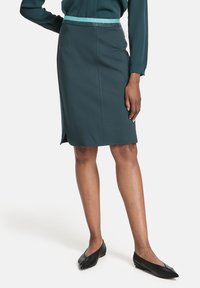 Kvinna som bär en mörk teal knälång kjol med ett ljusblått och mörk teal glittrande midjeband, kombinerat med svarta spetsiga flats.