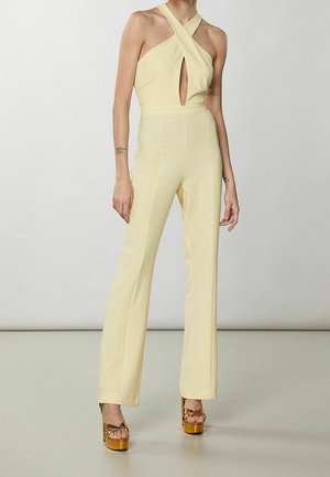 Mono amarillo con tirantes cruzados, detalle en forma de llave en el pecho, cuerpo ajustado y piernas acampanadas. Usado con tacones de plataforma dorados.