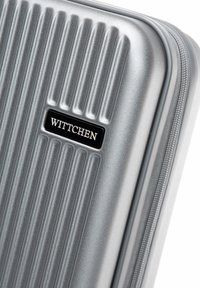 Valigia rigida argentata con scanalature verticali, dotata di una placca con il logo nero "WITTCHEN" e chiusura con zip.