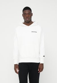 Junger Mann in einem weißen Kappa-Hoodie mit Vordertasche und schwarzen Hosen, der vor einem schlichten weißen Hintergrund steht.