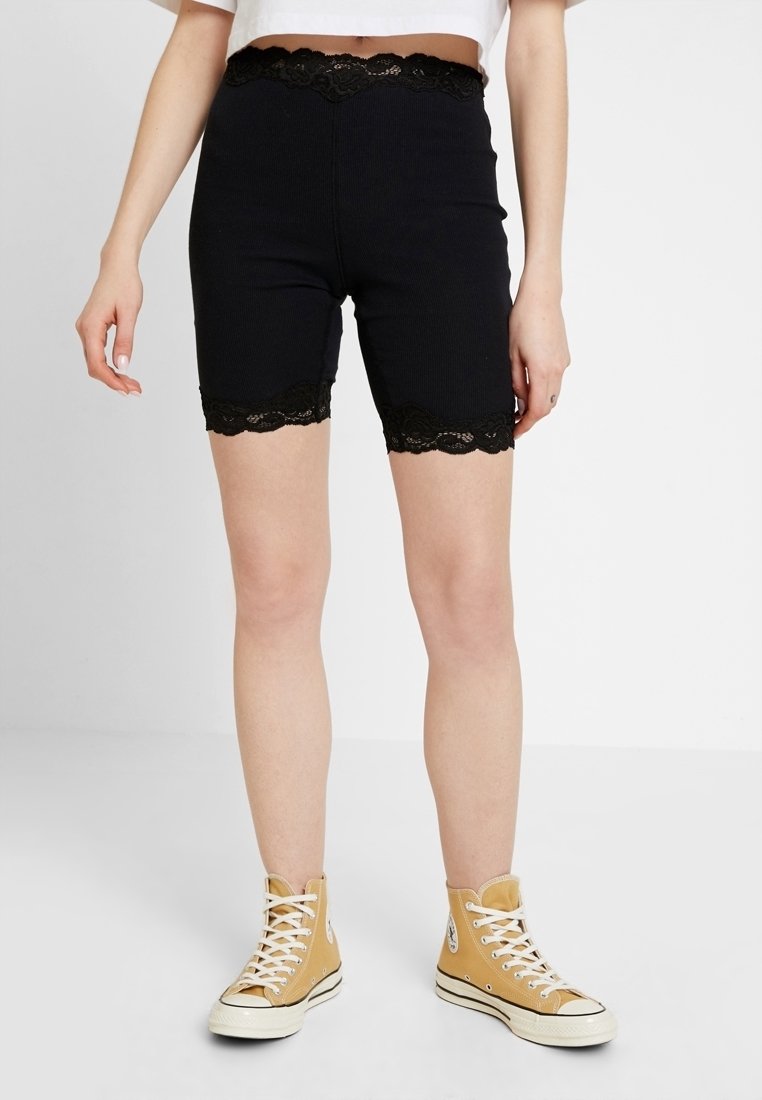 Svarta stretchshorts med spetsdetaljer vid midjan och fållen, med ribbad textur och figurnära design, bärs tillsammans med en vit topp och gula sneakers.