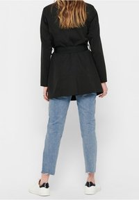 Schwarzer, hüftlanger Mantel mit einem Gürtel, kombiniert mit hellblauen, verwaschenen Jeans im geraden Schnitt und weißen Sneakers. Glattes Material, minimalistisches Design.