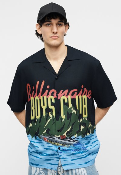 Νεαρός άνδρας φορώντας μαύρο καπέλο και μαύρο πουκάμισο με κουμπιά, με το κείμενο "Billionaire Boys Club" και γραφικό μιας βάρκας στο νερό.