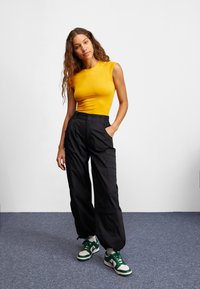 Gul tajt linne, svarta cargo-byxor med sidofickor och vita sneakers med gröna detaljer. Modellen står mot en blå matta som bakgrund.