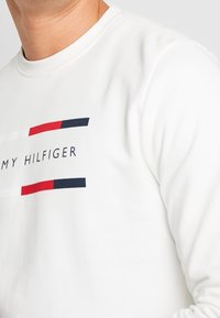 Bílá košile s dlouhým rukávem s detailním pohledem na trup a krk, se červeným, bílým a modrým logem Tommy Hilfiger přes hruď.