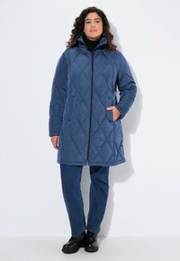 Vadderad blå pufferjacka med hög krage, framdragkedja och sidofickor. Bärs över svart polotröja och blå jeans, avslutat med svarta stövlar.