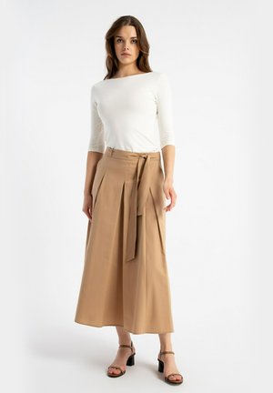 Kobieta stojąca, ubrana w biały top z rękawem do łokcia, beżowe szerokie spodnie z wysokim stanem i paskiem oraz brązowe sandały na słupku.
