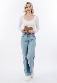 Frau mit blondem Haar, die ein weißes Oberteil und blaue Jeans trägt, hält eine braune Geldbörse und steht vor einem einfarbigen weißen Hintergrund.