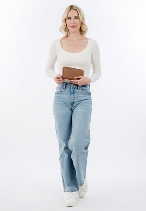 Femme aux cheveux blonds portant un haut blanc et un jean bleu tenant un portefeuille marron, debout devant un fond blanc uni.