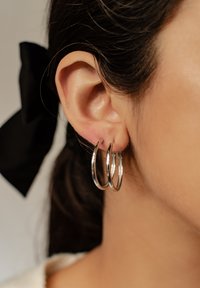 Pendientes de aro de plata que presentan tres bucles interconectados. Textura suave con un acabado brillante, situados frente a un fondo de cabello oscuro.