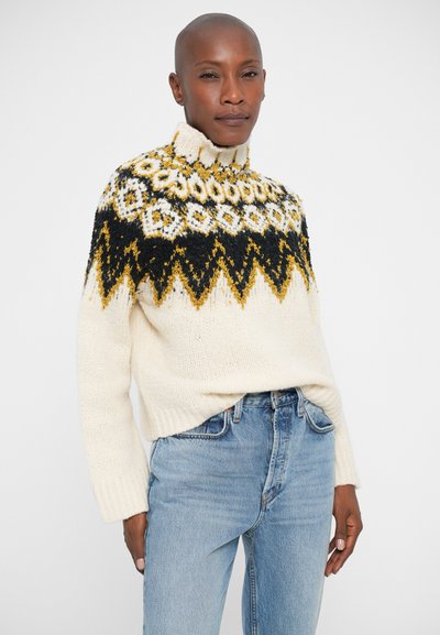 Persona che indossa un maglione a collo alto crema con motivi geometrici neri e gialli, abbinato a jeans a vita alta di un blu chiaro.