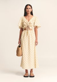 Dvodelna obleka z rumenimi pikami, ki vključuje kratko zgornjo majico z zavezovanjem spredaj in dolgo krilo, izdelano iz lahkega blaga. Doplačana s tkanim torbico.