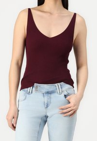 Débardeur côtelé bordeaux avec un décolleté en V profond, associé à un jean en denim bleu clair, doté de poches boutonnées et d'une coupe ajustée.