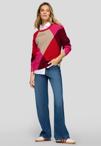 Bunter gestrickter Pullover mit Rautenmuster in Rot, Pink und Beige; kombiniert mit blauen Schlagjeans und weißen Turnschuhen.