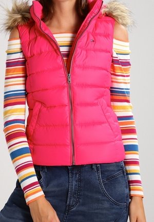 Pink gesteppte ärmellose Jacke mit pelzbesetzter Kapuze, getragen über einem Cold-Shoulder-Oberteil mit mehrfarbigen horizontalen Streifen und dunklen Jeans.