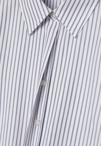 Chemise blanche à boutons avec rayures verticales bleues. Elle présente un col classique, des boutons opaques et une texture douce en coton.