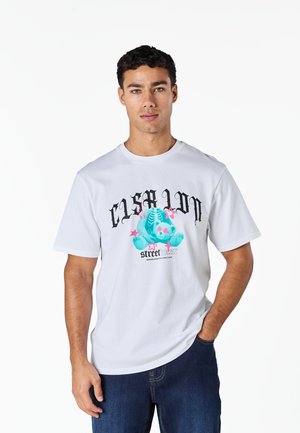 Mann trägt weißes T-Shirt mit türkisem Skelettbären und pinken Sternen, kombiniert mit dunkelblauer Jeans, steht vor weißem Hintergrund.