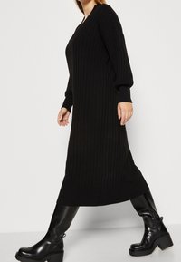 Robe longue noire à manches longues en tissu côtelé avec un col en V, présentant une coupe décontractée et un tissu texturé, associée à des bottines noires brillantes.
