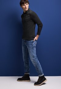 Czarna bluzka z długim rękawem o teksturowym wykończeniu i kieszonką na piersi, w połączeniu z jasnoniebieskimi dżinsami oraz czarnymi botkami na kostkę z grubą podeszwą.