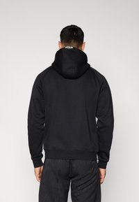 Sweat à capuche noir avec cordon de serrage, poignets et ourlet côtelés. Présente un logo dans le dos et un tissu lisse et texturé.