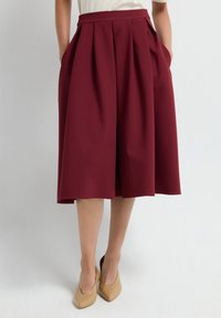 Jupe plissée bordeaux avec poches latérales, fabriquée en tissu texturé, arrivant au genou, portée avec des chaussures à talons hauts nude.