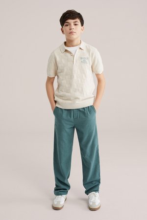 Fiú áll zsebre tett kézzel, krémszínű kockás pólót és türkiz nadrágot visel fehér tornacipővel, sima bézs háttér előtt.