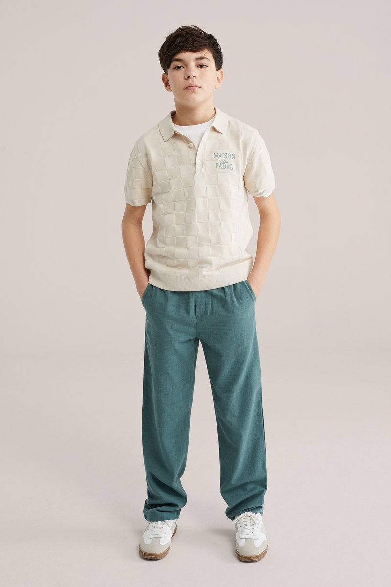 Chlapec stojí s rukami v vreckách, oblečený v krémovej károvanej polokošeli a tyrkysových nohaviciach s bielymi teniskami, na jednoduchom béžovom pozadí.