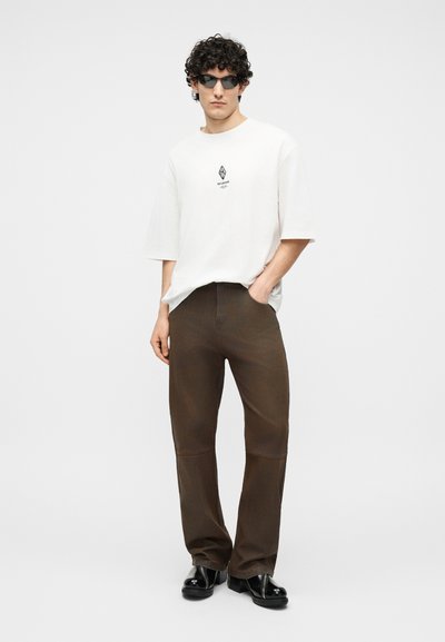 Άνδρας με σγουρά μαλλιά που φοράει μαύρα γυαλιά ηλίου, λευκό μεγάλο t-shirt με λογότυπο, καφέ παντελόνι και μαύρα γυαλιστερά παπούτσια, στέκεται με το ένα χέρι στην τσέπη.