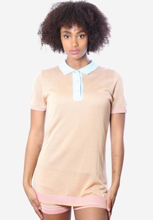 Junge Frau mit lockigem Haar, die ein beiges kurzärmeliges Poloshirt und pinke Shorts trägt, vor einem schlichten weißen Hintergrund stehend.